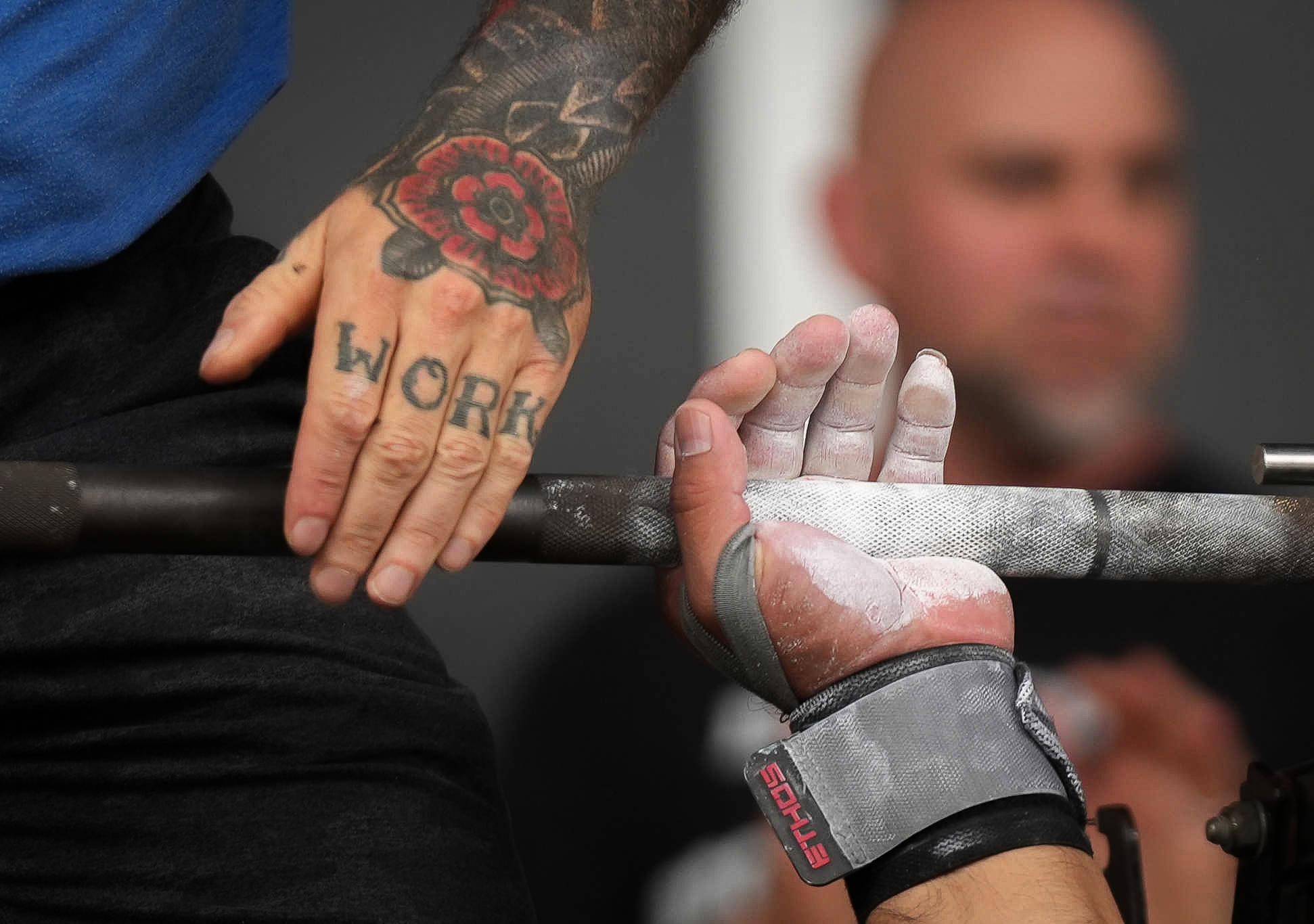 Close up of a person benchpressing with a powdered hand, with support from a person with a hand with a tattoo that says "work"