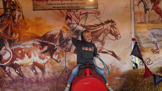 Young boy holding a lasso and riding on a red mechanical bull.