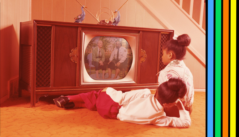 Two children sit on the floor in front of a console television watching Mister Rogers