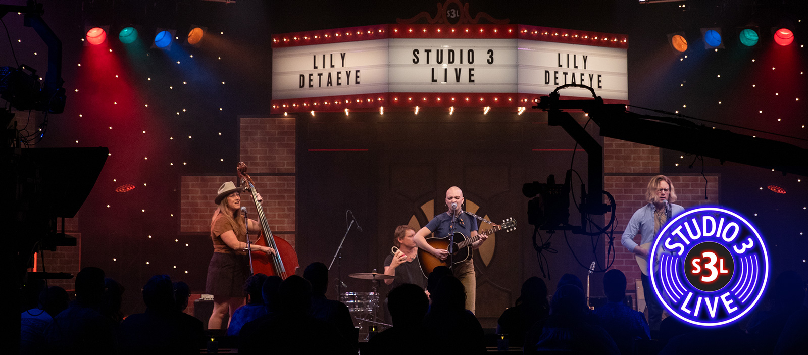 A white woman sings and plays guitar on stage under a lit Studio 3 LIVE marquee