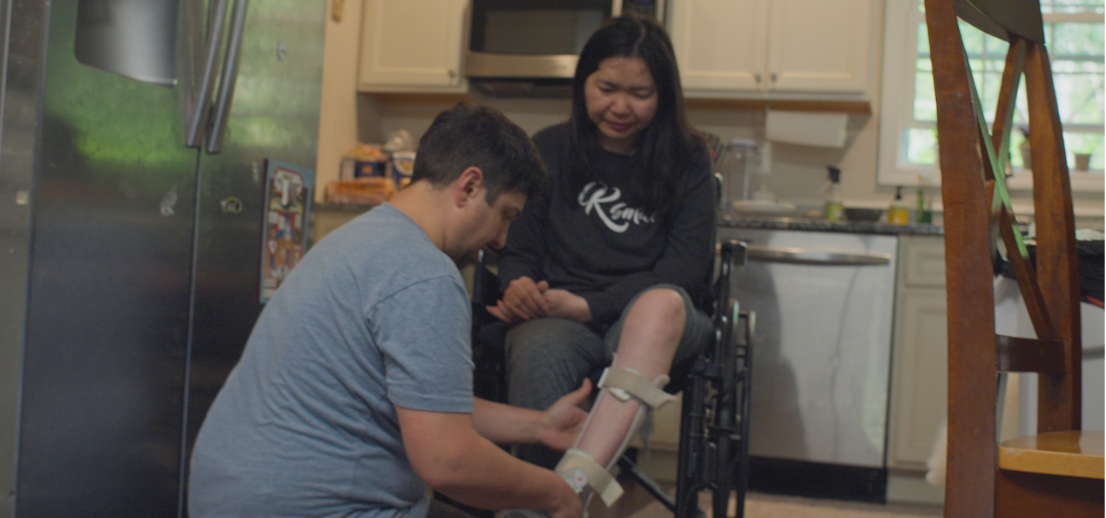 A person adjusts a leg brace for another person in a wheelchair in a kitchen.