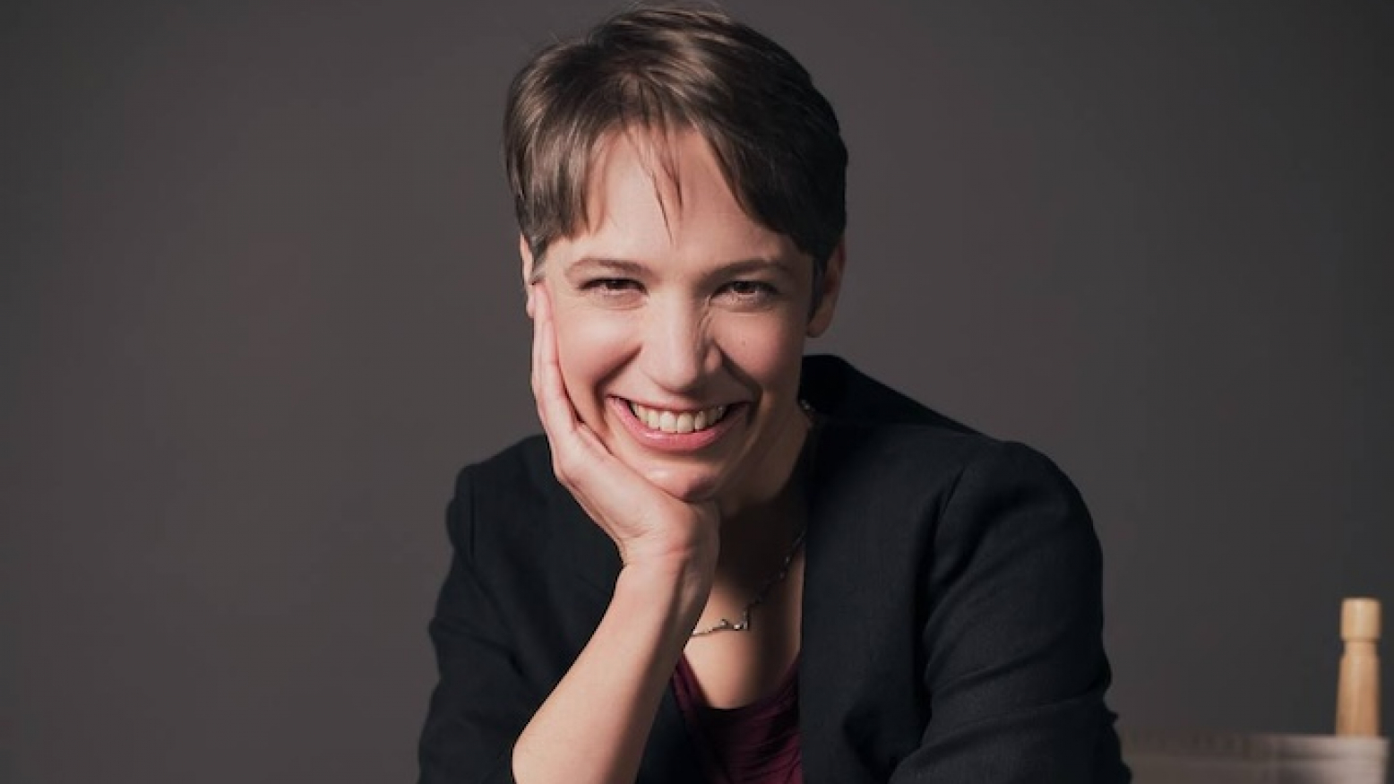 Charity Nebbe, a white woman with short brown hair wearing a black blazer, smiles with her chin resting in her hand. 