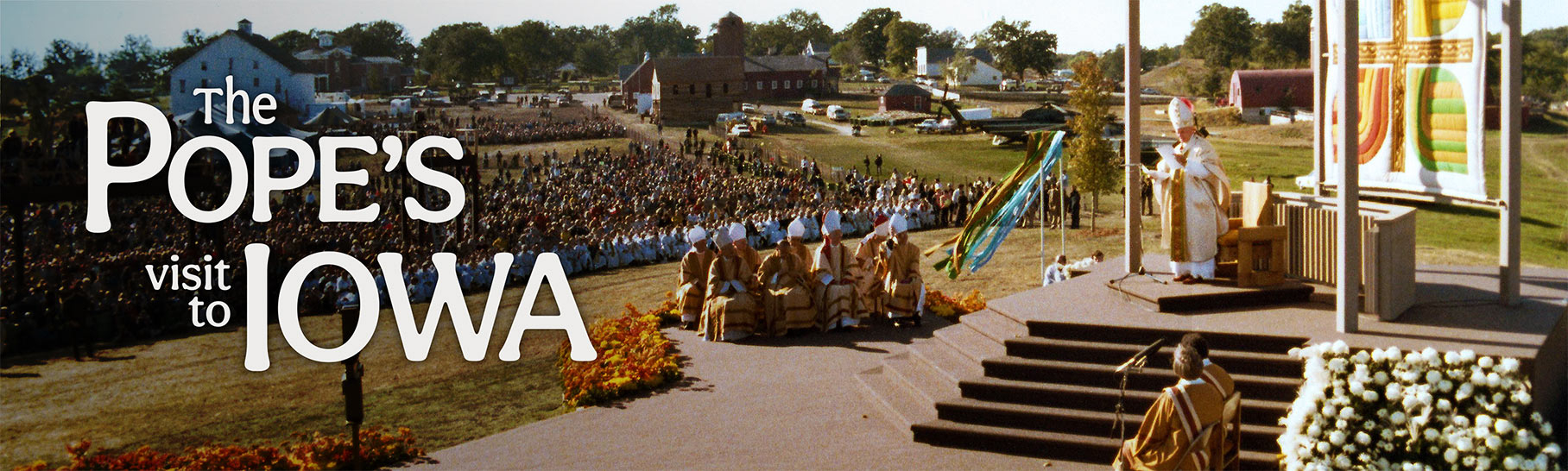 The Pope's Visit to Iowa, a person wearing a long white robe and tall, diamond shaped, ornamental hat stands on a small stage before a large crowd.