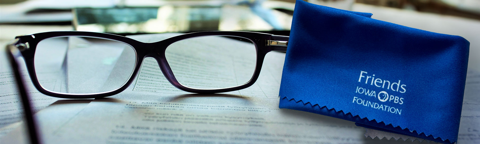 Reading glasses sit on an open book with a royal blue lens cloth with the Friends Iowa PBS Foundation logo