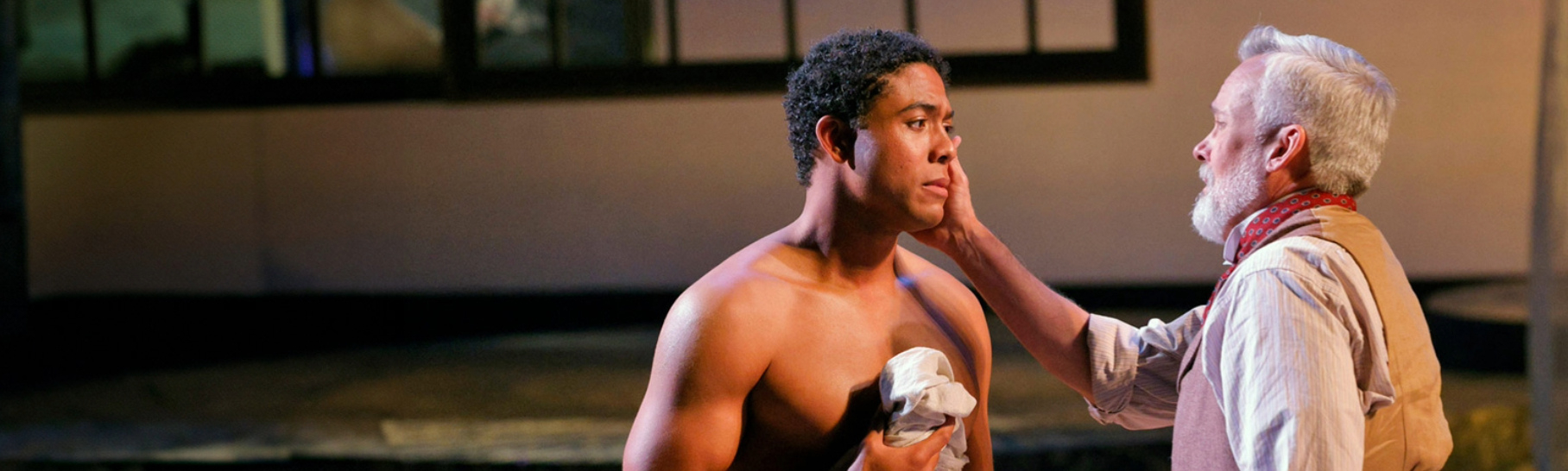 A powerful scene from Des Moines Metro Opera's production of "American Apollo." A shirtless young man, portraying Thomas Eugene McKeller, stands with a somber expression as an older man, playing artist John Singer Sargent, gently touches his face in a tender and emotional moment.