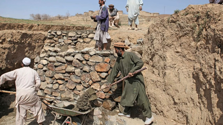 Men dressed in tunics shovel rock and dirt into a wheelbarrow.