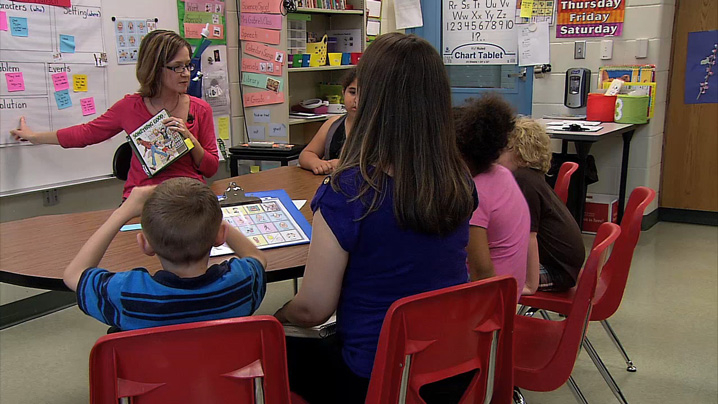 Five students sit at a table while a teacher points to the white board.