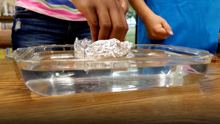 Two children test a boat made of aluminum foil in a dish of water.