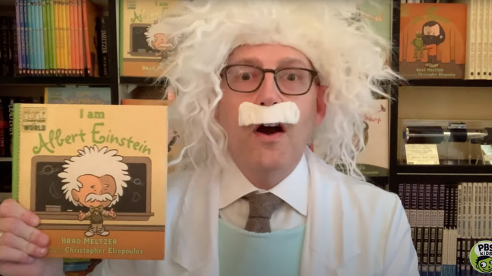 A man dressed in a white coat, white crazy-haired wig and white fake mustache holds up the book "I am Albert Einstein."