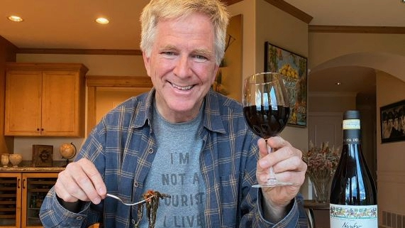 Rick Steves holding a fork eating dinner with a glass of red wine in the other hand.