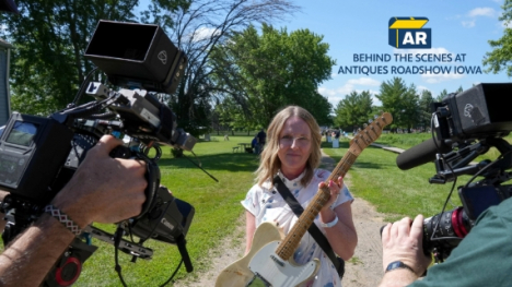 A woman holding a guitar with two people holding cameras filming her