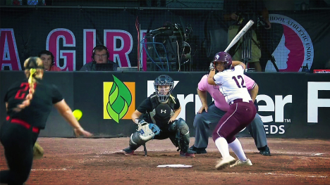 An IGHSAU Girls State Softball Championship game - a pitcher sends a ball towards the batter, catcher and umpire near home plate - members of the media in the background.