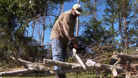 A Texas man in a brown work coat using a chainsaw to cut a downed tree into smaller parts for disposal.